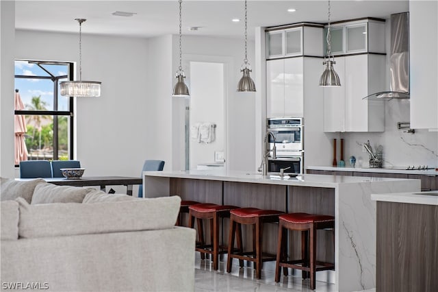 kitchen featuring wall chimney exhaust hood, stainless steel double oven, pendant lighting, a kitchen bar, and white cabinets