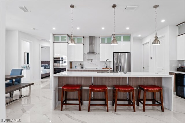 kitchen featuring a large island with sink, a kitchen breakfast bar, wall chimney exhaust hood, appliances with stainless steel finishes, and white cabinetry