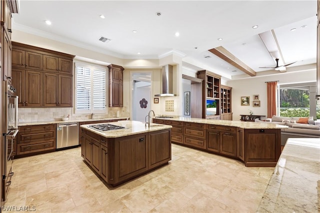 kitchen with appliances with stainless steel finishes, tasteful backsplash, a kitchen island with sink, light stone counters, and wall chimney range hood