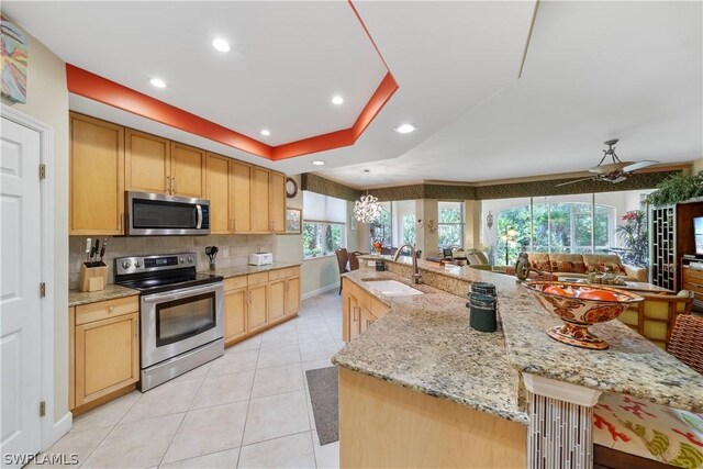 kitchen featuring a tray ceiling, stainless steel appliances, ceiling fan with notable chandelier, sink, and backsplash