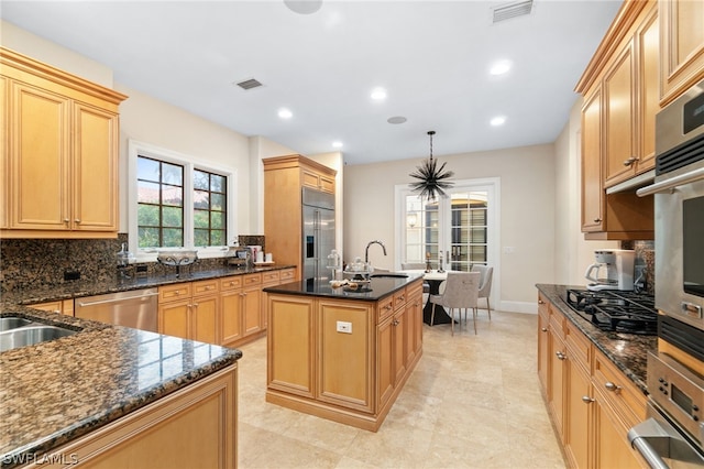 kitchen with sink, dark stone counters, pendant lighting, stainless steel appliances, and a kitchen island with sink