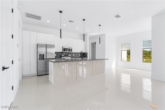 kitchen featuring tasteful backsplash, stainless steel appliances, white cabinets, hanging light fixtures, and an island with sink