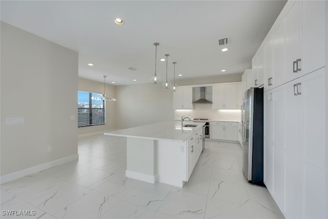 kitchen featuring wall chimney exhaust hood, decorative light fixtures, white cabinets, an island with sink, and stainless steel refrigerator