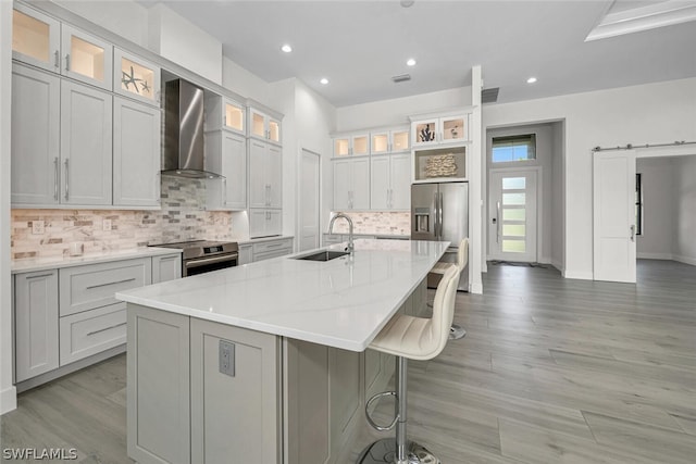 kitchen with wall chimney exhaust hood, stainless steel appliances, a center island with sink, sink, and a barn door