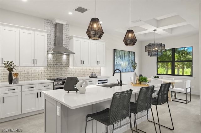 kitchen with backsplash, an island with sink, coffered ceiling, wall chimney exhaust hood, and sink