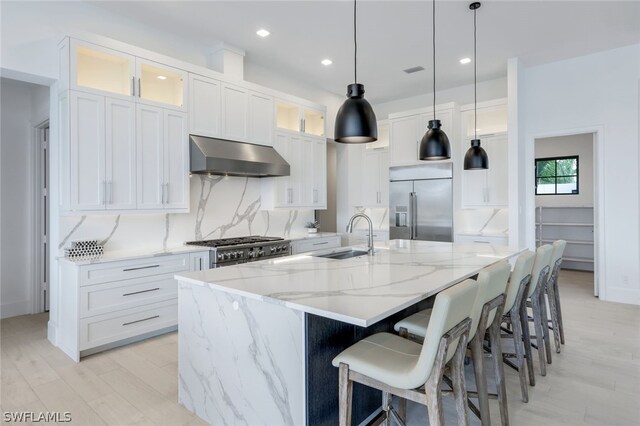 kitchen featuring sink, high quality appliances, light wood-type flooring, wall chimney exhaust hood, and a large island with sink