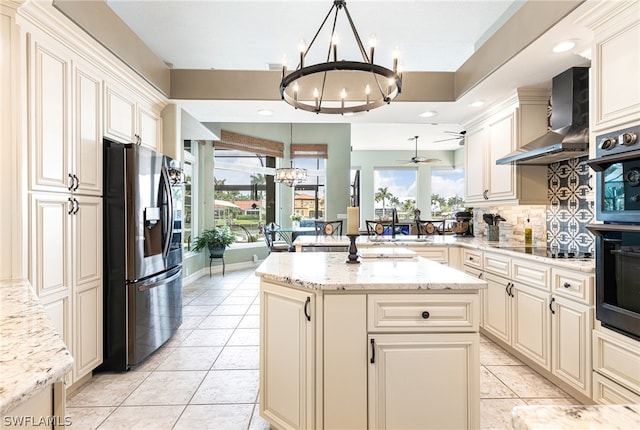 kitchen with pendant lighting, wall chimney range hood, stainless steel refrigerator with ice dispenser, light stone counters, and cream cabinetry