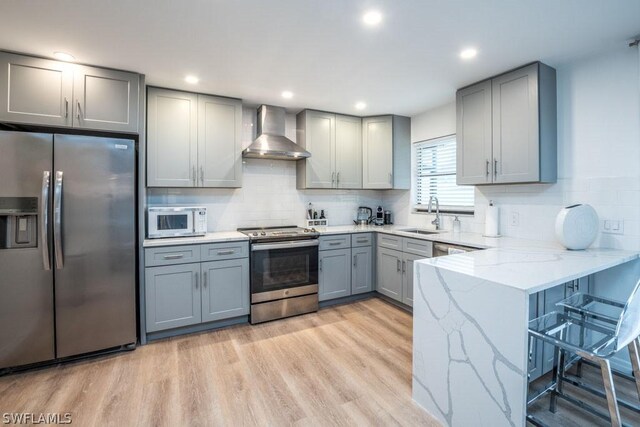 kitchen featuring kitchen peninsula, appliances with stainless steel finishes, light wood-type flooring, light stone counters, and wall chimney exhaust hood