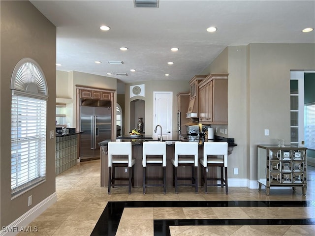 kitchen featuring a breakfast bar, a center island with sink, stainless steel built in fridge, kitchen peninsula, and range hood