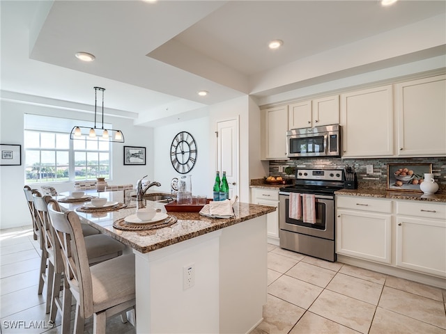 kitchen with appliances with stainless steel finishes, stone counters, light tile patterned floors, a kitchen island with sink, and a raised ceiling