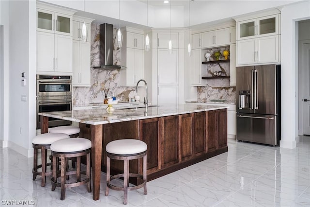 kitchen featuring appliances with stainless steel finishes, an island with sink, hanging light fixtures, light stone countertops, and wall chimney range hood