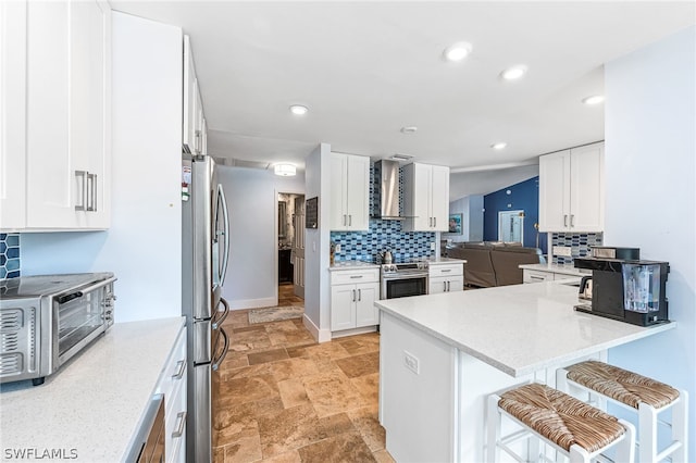 kitchen featuring wall chimney exhaust hood, a kitchen breakfast bar, kitchen peninsula, stainless steel appliances, and white cabinets