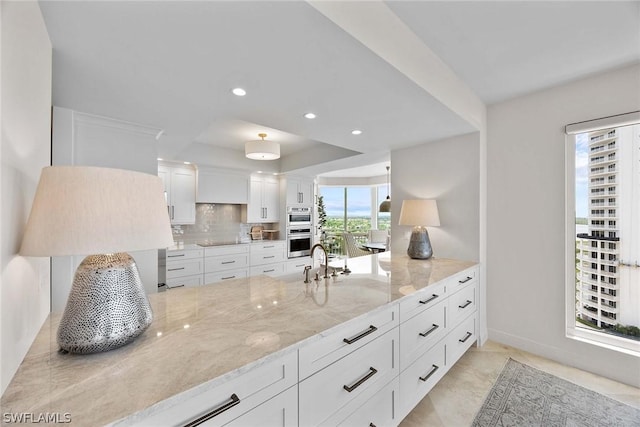 kitchen featuring white cabinetry, double oven, a tray ceiling, light stone countertops, and decorative backsplash