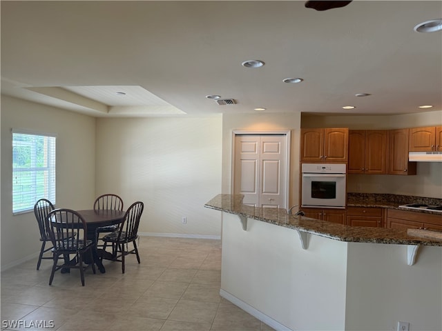 kitchen featuring dark stone countertops, light tile patterned floors, a raised ceiling, oven, and a kitchen bar