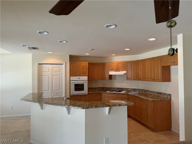 kitchen with white appliances, light tile patterned floors, a kitchen breakfast bar, an island with sink, and dark stone countertops