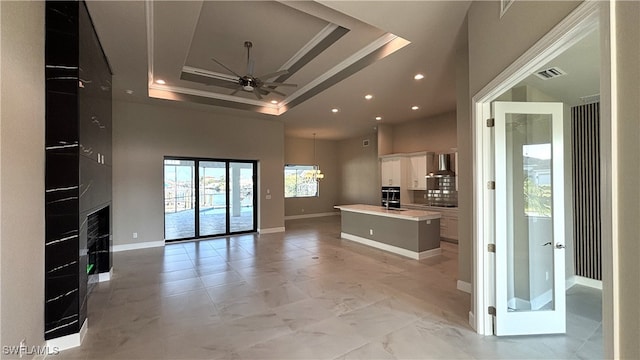 kitchen featuring wall chimney range hood, ceiling fan, white cabinetry, an island with sink, and a raised ceiling