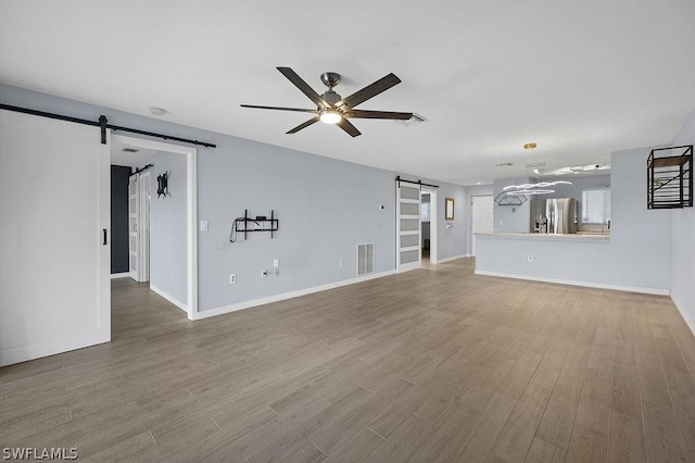 unfurnished living room featuring a barn door, wood finished floors, visible vents, a ceiling fan, and baseboards