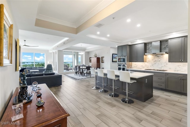 kitchen with ceiling fan, a kitchen island with sink, wall chimney range hood, a kitchen breakfast bar, and light hardwood / wood-style floors