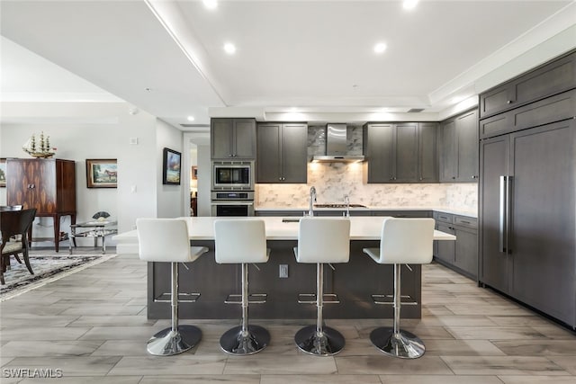 kitchen with a kitchen island with sink, wall chimney range hood, built in appliances, and a breakfast bar area