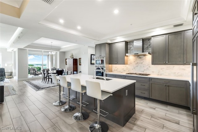 kitchen with a kitchen island with sink, wall chimney exhaust hood, a tray ceiling, stainless steel appliances, and a breakfast bar area