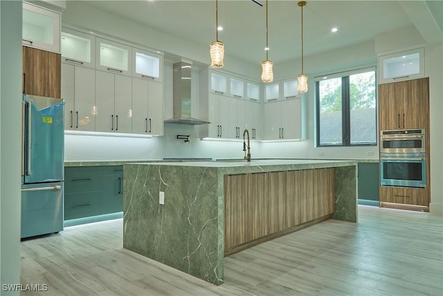 kitchen featuring glass insert cabinets, appliances with stainless steel finishes, white cabinetry, a large island with sink, and wall chimney exhaust hood