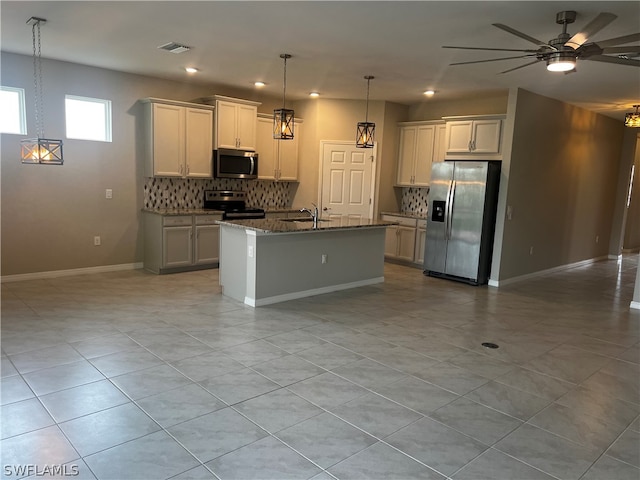 kitchen with ceiling fan, appliances with stainless steel finishes, a kitchen island with sink, backsplash, and dark stone counters