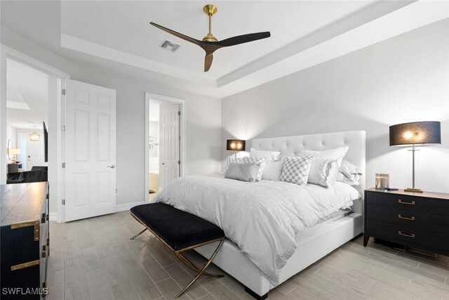 bedroom featuring wood finish floors, a raised ceiling, visible vents, ensuite bath, and baseboards