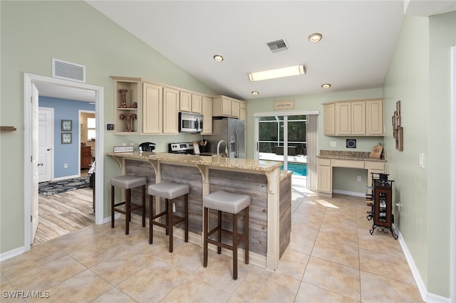 kitchen with cream cabinets, vaulted ceiling, light stone countertops, appliances with stainless steel finishes, and a kitchen bar