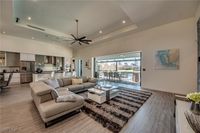 living room featuring recessed lighting, a towering ceiling, baseboards, light wood-style floors, and a tray ceiling