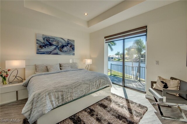 bedroom featuring access to exterior, hardwood / wood-style floors, a water view, and a raised ceiling