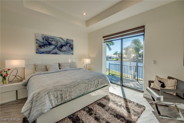 bedroom with recessed lighting, a water view, wood finished floors, access to outside, and a tray ceiling