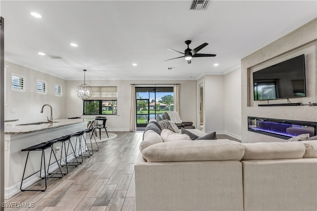 living room with ceiling fan with notable chandelier, light hardwood / wood-style flooring, crown molding, and sink