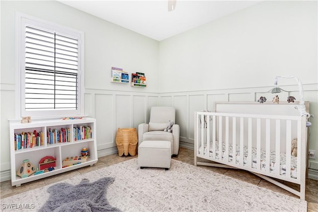 tiled bedroom featuring a decorative wall, a nursery area, a wainscoted wall, and ceiling fan