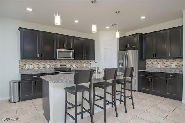 kitchen with appliances with stainless steel finishes, an island with sink, hanging light fixtures, light tile patterned floors, and light stone counters