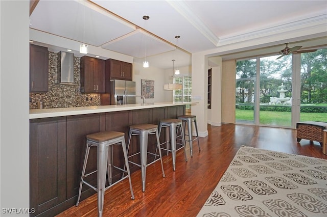 kitchen with wall chimney exhaust hood, stainless steel refrigerator with ice dispenser, a kitchen bar, decorative backsplash, and ceiling fan