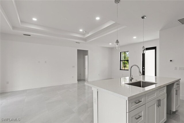 kitchen with a raised ceiling, an island with sink, white cabinetry, sink, and decorative light fixtures