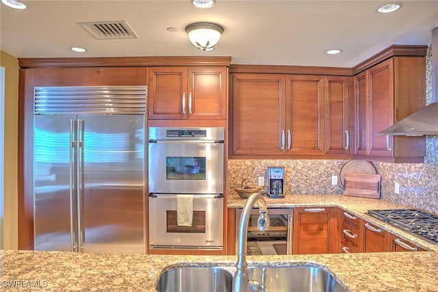 kitchen featuring decorative backsplash, light stone countertops, wall chimney exhaust hood, stainless steel appliances, and sink