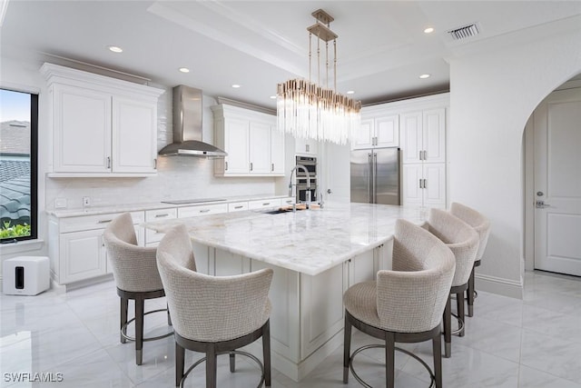 kitchen featuring wall chimney range hood, a tray ceiling, an island with sink, stainless steel appliances, and white cabinets