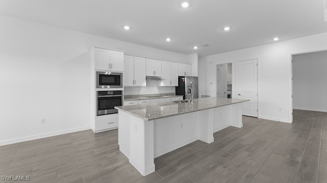 kitchen featuring appliances with stainless steel finishes, a kitchen island with sink, white cabinetry, light stone countertops, and hardwood / wood-style flooring