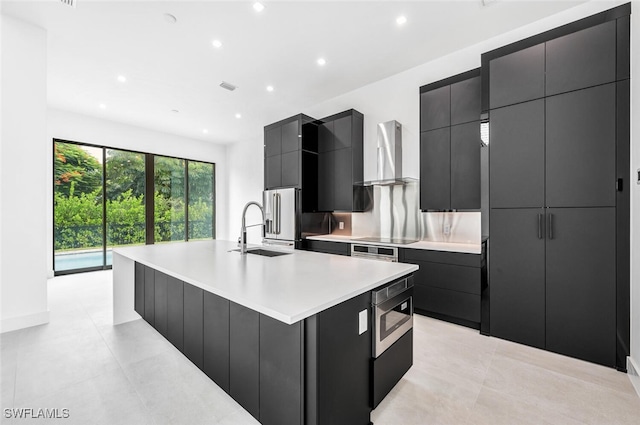 kitchen with appliances with stainless steel finishes, a sink, dark cabinetry, a large island with sink, and wall chimney exhaust hood