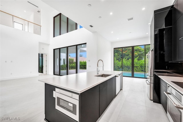 kitchen featuring appliances with stainless steel finishes, a towering ceiling, sink, light tile patterned floors, and an island with sink