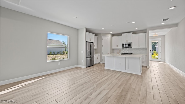 kitchen with appliances with stainless steel finishes, backsplash, light wood-type flooring, an island with sink, and white cabinetry