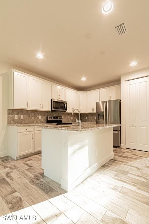 kitchen featuring white cabinets, decorative backsplash, an island with sink, appliances with stainless steel finishes, and light stone counters