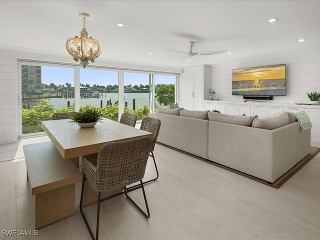 dining area with ceiling fan with notable chandelier, a water view, light hardwood / wood-style flooring, and wooden walls