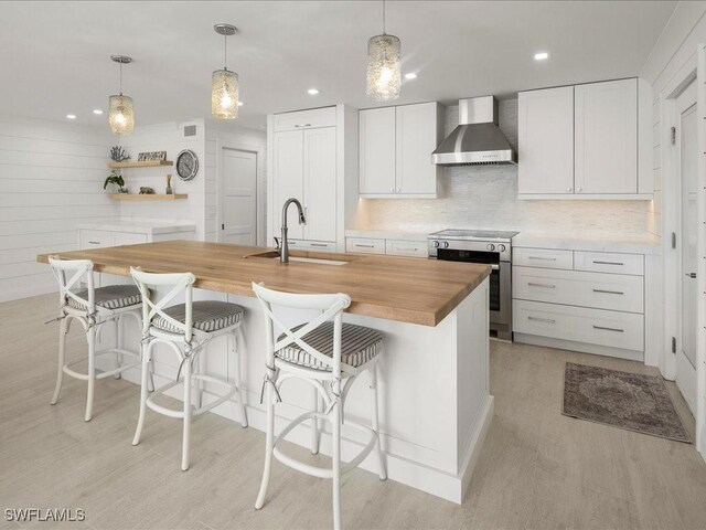 kitchen with wooden counters, a center island with sink, sink, wall chimney exhaust hood, and white cabinetry