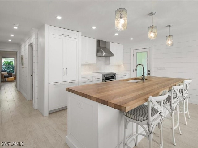 kitchen with wall chimney exhaust hood, decorative light fixtures, a center island with sink, light hardwood / wood-style floors, and white cabinetry