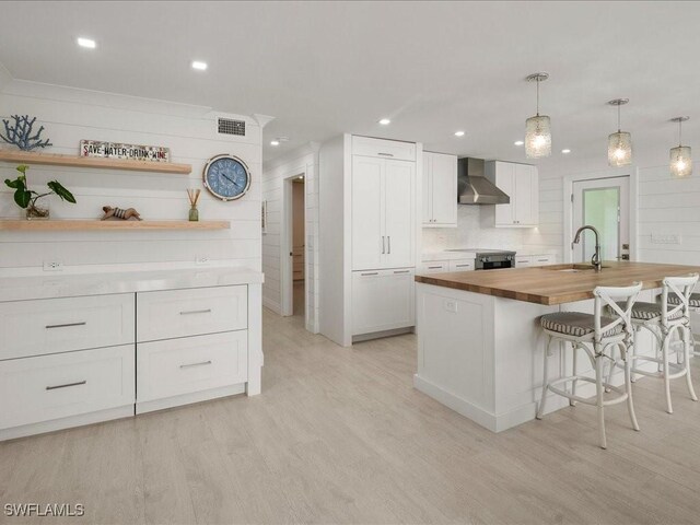 kitchen with white cabinets, butcher block countertops, a kitchen island with sink, and wall chimney exhaust hood