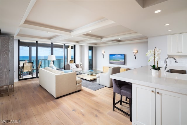 living room featuring light wood-type flooring, a wall of windows, a water view, coffered ceiling, and sink