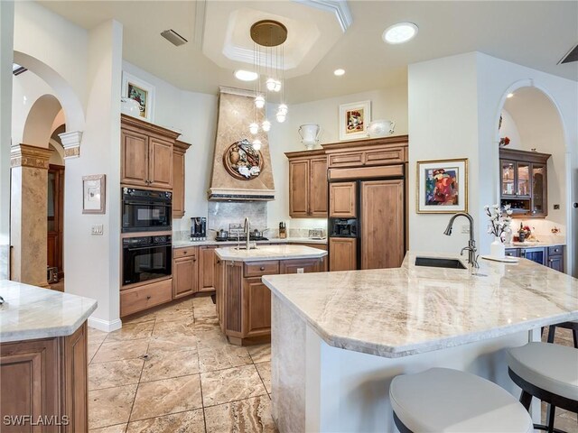 kitchen with sink, black double oven, a kitchen island with sink, a raised ceiling, and light stone countertops