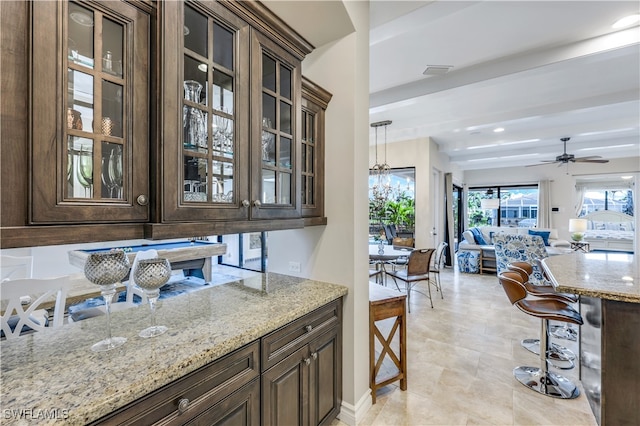 kitchen featuring ceiling fan with notable chandelier, dark brown cabinets, pendant lighting, light stone countertops, and a breakfast bar area